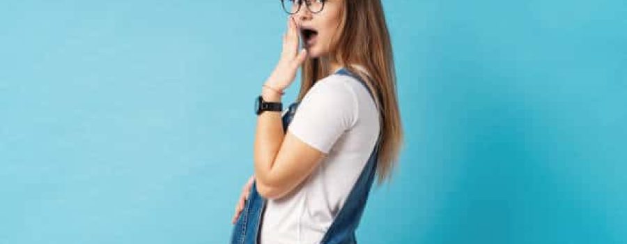 Portrait of good-looking young, cheerful pregnant woman wearing comfy denim romper and glasses making surprised face, looking at camera with a happy face expression over blue background.